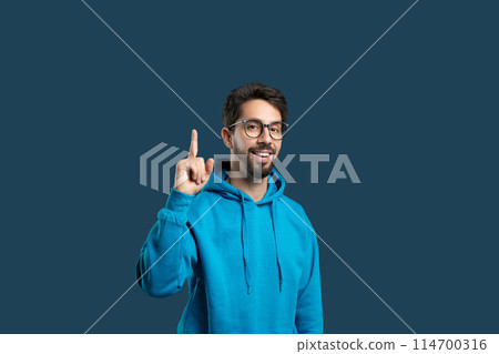 A cheerful young man with glasses and a beard is dressed in a casual blue hoodie, lifting his finger to point above his head, expressing an idea or solution 114700316