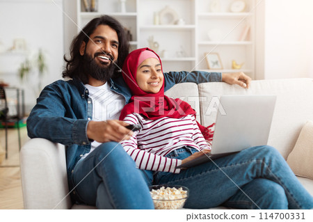 Muslim couple seated comfortably on a couch, focusing on a laptop placed in front of them, watching TV. They appear engaged in an online activity or work 114700331