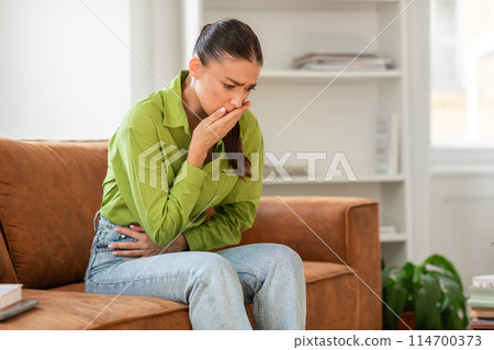 A woman clad in a green shirt and jeans is sitting on a brown sofa, looking distressed with her hand over her mouth and clutching her stomach, a possible sign of nausea or abdominal discomfort A woman clad in a green shirt and jeans is sitting on a brown sofa, looking distressed with her hand over her mouth and clutching her stomach, a possible sign of nausea or abdominal discomfort 114700373