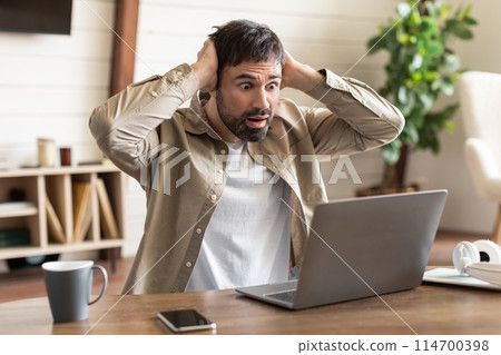 A man is seated at a wood table, hands on his head with an expression of shock and disbelief as he looks at his laptop screen. 114700398