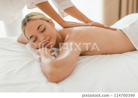 The image captures a serene moment of a blonde young woman getting a soothing back massage at a spa, highlighting her relaxed facial expression 114700399