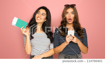 Two young women with similar striped shirts stand side-by-side against a vibrant pink backdrop, holding smartphone and passport with tickets. Hot traveling offer Two young women with similar striped shirts stand side-by-side against a vibrant pink backdrop, holding smartphone and passport with tickets. Hot traveling offer 114700401
