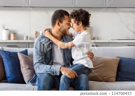African American man is seated on the armrest of a couch, with one arm wrapped protectively around a little boy who is sitting beside him. They appear to be engaged in a bonding moment. 114700457