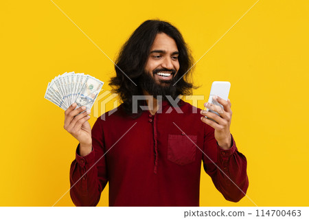Indian man with long hair and a beard holding cash and a cell phone in his hands. He seems to be engaging in a transaction or communication. His expression is focused and attentive. 114700463