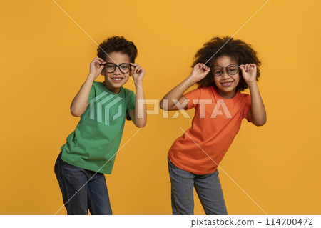 Two African American children, both wearing glasses, are striking a pose for a camera. They seem cheerful and excited, looking directly at the lens with big smiles Two African American children, both wearing glasses, are striking a pose for a camera. They seem cheerful and excited, looking directly at the lens with big smiles 114700472