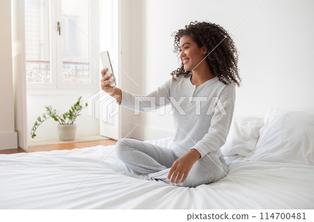A Hispanic woman is sitting on a bed, holding her phone up to take a selfie. She is smiling and looking at the screen of the phone as she captures the moment. 114700481