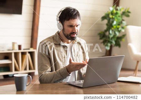 A man sitting with headphones on, focusing on a laptop screen. He appears engaged and attentive to the information displayed on the computer, have video call 114700482
