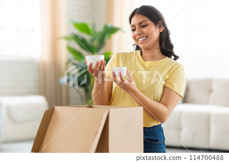 A cheerful young middle eastern woman is standing in a sunlit living room, holding and examining two items she has just unpacked from a cardboard box 114700488
