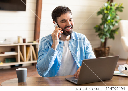 A man wearing a blue shirt is sitting at a wooden table, engaged in a phone conversation on his cell phone. The man appears focused as he speaks into the phone, his expression serious. 114700494