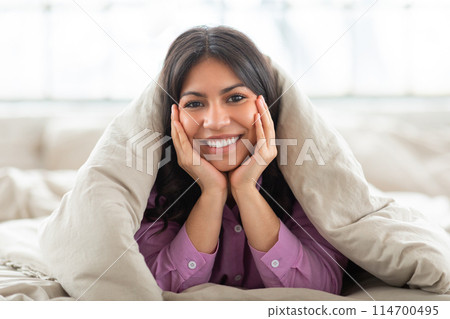 A middle eastern woman is lying on top of a bed, covered by a blanket. She appears comfortable and relaxed, resting peacefully indoors. 114700495