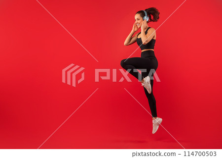 A joyful young woman with her hair tied in a ponytail is captured mid-jump against a bold red backdrop, showcasing her active lifestyle, using wireless headphones, copy space 114700503