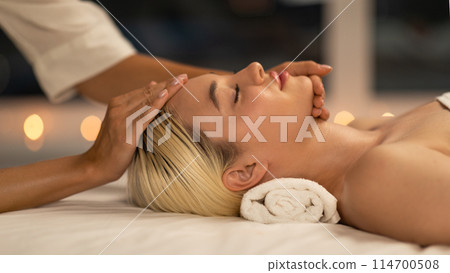 The photo shows a blonde woman lying down during a facial massage at a spa, with a calm and peaceful expression, enhancing her youthful glow The photo shows a blonde woman lying down during a facial massage at a spa, with a calm and peaceful expression, enhancing her youthful glow 114700508