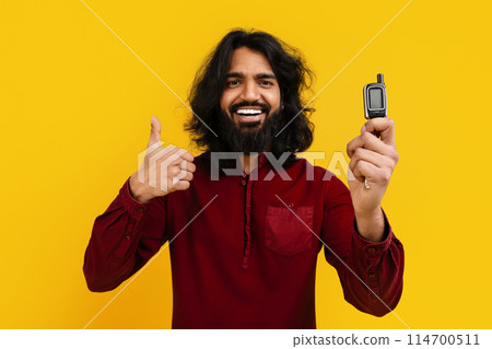 Cheerful happy excited vivid driver young bearded Indian man holding in hand new car key and showing thumb up, isolated on yellow background, studio shot, copy space. Driving, car leasing concept 114700511