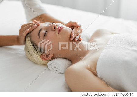 This image captures a blonde young woman peacefully enjoying a facial massage at a luxury spa, highlighting her relaxed facial expressions 114700514