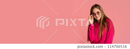 Pretty long-haired girl adjusting stylish sunglasses, posing isolated on pink studio background, smiling at camera, panorama with copy space 114700516