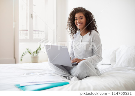 A Hispanic woman sits on a bed, focused on using a laptop computer. She types on the keyboard, with the screen displaying data. The room is well-lit, with a simple and clean decor. 114700521