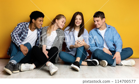 Four friends multiethnic teenagers are sitting closely together on the floor, leaning against a bright yellow wall. One of them is holding a smartphone, and they are all looking at the screen Four friends multiethnic teenagers are sitting closely together on the floor, leaning against a bright yellow wall. One of them is holding a smartphone, and they are all looking at the screen 114700577