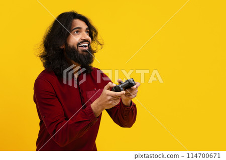 Indian man with a beard and long hair is standing against a bright yellow backdrop, wearing a red shirt and holding a game controller, copy space Indian man with a beard and long hair is standing against a bright yellow backdrop, wearing a red shirt and holding a game controller, copy space 114700671