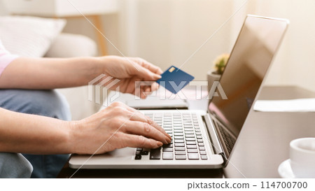 Lady holding a credit card in one hand while operating a laptop with the other hand, focused on making an online transaction, with computer for shopping or banking. 114700700