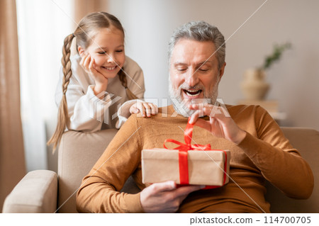 Senior man and a young girl are seated on a couch, both looking excitedly at a wrapped present between them. Grandfather is smiling, and the little girl appears curious. The room is cozy and well-lit. 114700705