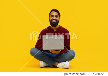 Indian man is seated on the floor, working on a laptop computer. He appears focused and engaged in his task, isolated on yellow background Indian man is seated on the floor, working on a laptop computer. He appears focused and engaged in his task, isolated on yellow background 114700720