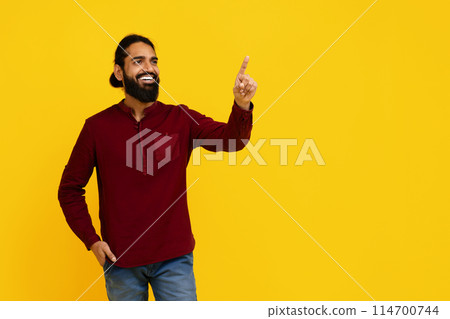 Indian cheerful man with a beard is standing against a bright yellow backdrop, pointing upwards with his left hand, touching invisible virtual screen Indian cheerful man with a beard is standing against a bright yellow backdrop, pointing upwards with his left hand, touching invisible virtual screen 114700744
