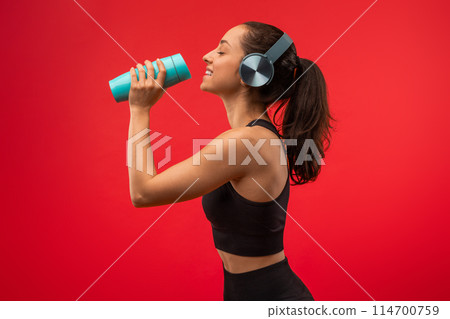 A woman wearing headphones is holding a blue cup to her lips, taking a sip of a beverage. She appears focused on enjoying her drink. 114700759