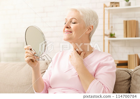 Senior woman is seated on a couch, holding a mirror in her hand. She appears to be examining her neck intently. The room is dimly lit, with soft furniture in the background. Senior woman is seated on a couch, holding a mirror in her hand. She appears to be examining her neck intently. The room is dimly lit, with soft furniture in the background. 114700784