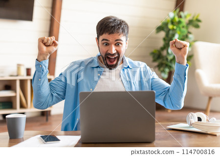 A jubilant young man raises his fists in triumphant celebration while sitting at a desk in a cozy home office setting. In front of him, an open laptop hints at the possible source of his excitement 114700816