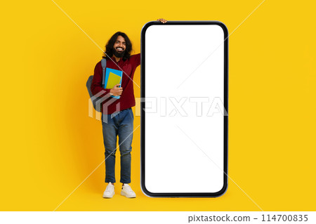 Cheerful Indian man with a beard student stands to the left side, holding colorful books and leaning against a massive, blank screen smartphone 114700835