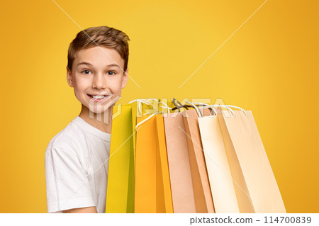 Cheerful teen boy holding lots of shopping bags and smiling, orange studio background Cheerful teen boy holding lots of shopping bags and smiling, orange studio background 114700839