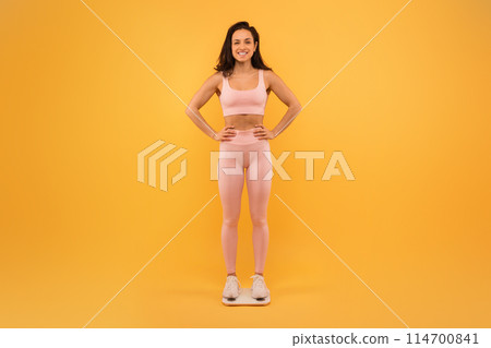 A smiling young woman stands confidently with her hands on her hips, dressed in a light pink sports bra and leggings, checking her weight 114700841
