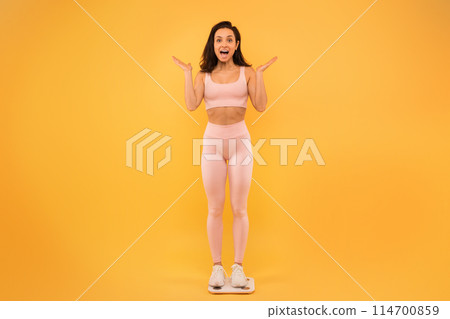 A young woman stands on a scale, expressing surprise and excitement with her hands raised. She is dressed in light-colored active wear and white sneakers, set against a vibrant yellow backdrop. 114700859
