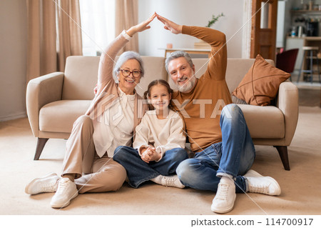 A smiling elderly couple and a young girl sit on the floor in a cozy living room, forming a house outline with their hands above them, symbolizing unity and family love. 114700917