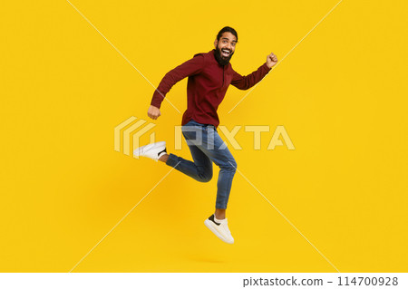 Indian man wearing a red sweater is captured mid-jump in the air. His arms are raised, and his legs are bent as he springs upwards. The background is yellow, emphasizing the mans movement. 114700928