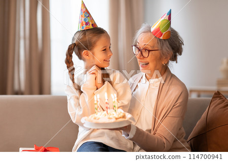 A cheerful grandmother wearing a party hat is presenting a birthday cake with lit candles to a young girl, also in a party hat, as they share a loving glance and a warm A cheerful grandmother wearing a party hat is presenting a birthday cake with lit candles to a young girl, also in a party hat, as they share a loving glance and a warm 114700941
