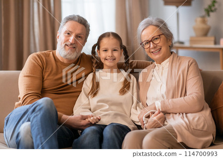 A heartwarming scene captures a young girl seated between her grandparents, sharing a moment of joy at home interior A heartwarming scene captures a young girl seated between her grandparents, sharing a moment of joy at home interior 114700993