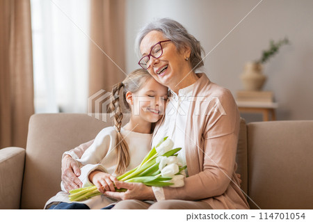 A cheerful elderly woman with glasses is warmly hugging her young granddaughter, who holds a bouquet of fresh white tulips, home interior 114701054