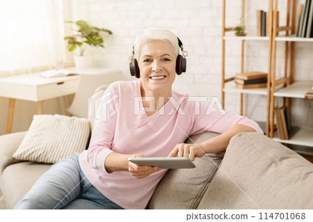 Senior woman enjoying music and watching videos on tablet pc in headphones, free space 114701068