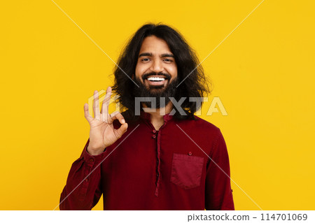 Indian man with a full beard and long hair smiles broadly, standing against a vivid yellow background, showing OK gesture with his right hand, expressing positivity and approval. 114701069