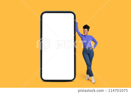 Hispanic woman stands next to a large smartphone with white blank screen isolated on yellow background, mockup copy space 114701075
