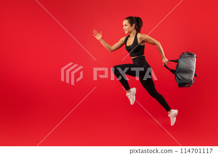A young woman is captured mid-jump against a vibrant red backdrop, exuding energy and happiness. She is dressed in sporty black attire and white sneakers, with a backpack in one hand A young woman is captured mid-jump against a vibrant red backdrop, exuding energy and happiness. She is dressed in sporty black attire and white sneakers, with a backpack in one hand 114701117