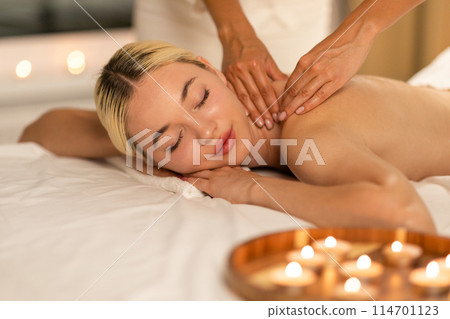 Beautiful woman lies face down on a massage table, receiving a back massage from a professional masseuse in a spa setting. The masseuses hands apply pressure on the woman back Beautiful woman lies face down on a massage table, receiving a back massage from a professional masseuse in a spa setting. The masseuses hands apply pressure on the woman back 114701123