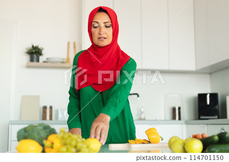A woman wearing a red and green hijab is shown in the act of chopping a variety of vegetables on a cutting board. 114701150