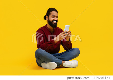 Indian man is seated on the floor, engrossed in his cell phone. He appears focused as he scrolls through the screen, possibly engaging in messaging or browsing 114701157