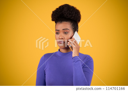 Hispanic woman is having a phone conversation while wearing a purple shirt. She is standing outdoors, holding the phone to her ear and speaking. 114701166