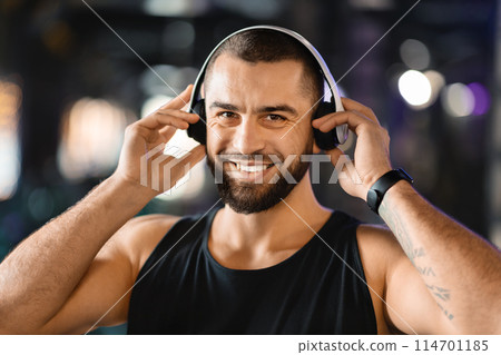 A man with a beard is wearing headphones, listening to music or audio. He appears focused or relaxed as he enjoys workout at gym, closeup 114701185