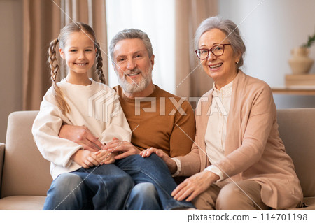 A heartwarming scene captures a young girl with long, braided hair sitting between her grandparents on a beige couch. The elderly couple and the girl are all smiling warmly A heartwarming scene captures a young girl with long, braided hair sitting between her grandparents on a beige couch. The elderly couple and the girl are all smiling warmly 114701198