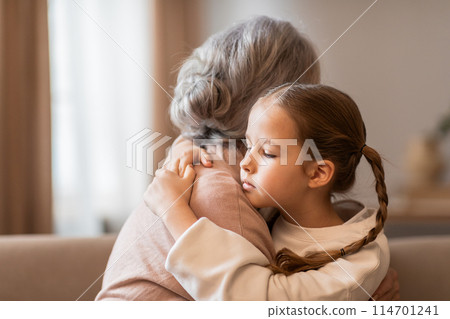 A young girl embraces a senior woman warmly as they sit together on a couch in a cozy living room. The child arms are wrapped around the older woman 114701241