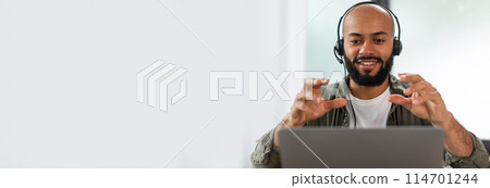Engaged black man wearing headphones while gesturing during a video call, suitable for web-banner with ample copy space Engaged black man wearing headphones while gesturing during a video call, suitable for web-banner with ample copy space 114701244
